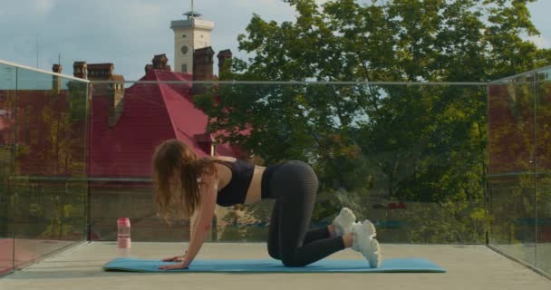 En sportig tjej gör ett benlyft medan hon knäböjer. Hon gör en övning på en yogamatta och det finns en flaska vatten bredvid henne. 4K — Stockvideo