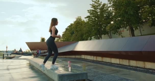 Una chica deportiva está haciendo una subida al banco en una pierna. Hay una botella de agua en el banco. Lleva leggins y gafas de sol. 4K — Vídeos de Stock