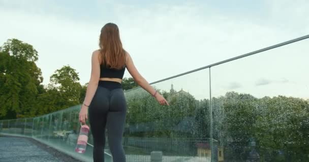 Ein sportliches Mädchen läuft mit dem Finger am gläsernen Geländer entlang. Sie trägt Leggings und hält eine Wasserflasche in der Hand. Schießen von hinten. 4K — Stockvideo