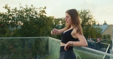 Spor kızı cam korkuluklara yaslanıyor. Akıllı seğirme ile ilgili eğitim bilgilerini inceliyor. Spor üniforması ve güneş gözlüğü giyiyor. Yakın plan çekim. 4K