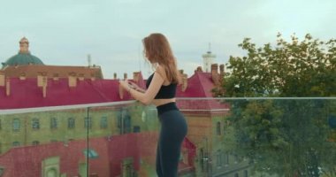 Sportif bir kız ayakta ve telefonda mesaj atıyor. O, cam korkuluklara yaslanıyor. Kız spor üniforması ve pembe gözlük takıyor. 4K