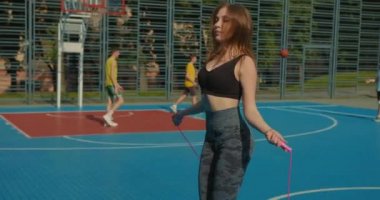 Sportif bir kız pembe atlama ipinden atlıyor. Saçları uçuyor. O bir spor üniforması giyiyor. Arka planda insanlar basketbol oynuyor. 4K