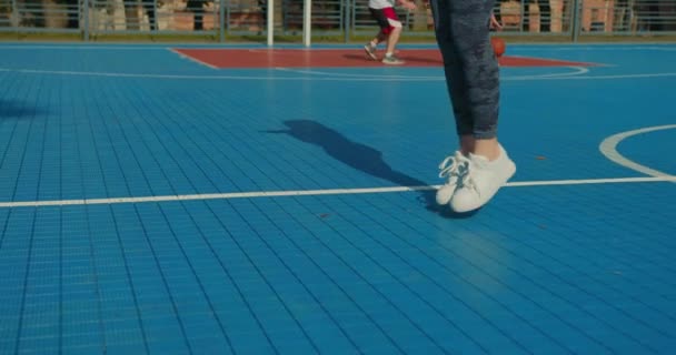 Disparos de cerca. Una chica deportiva está saltando una cuerda rosa. Su pelo está volando. Se está vistiendo con uniforme deportivo. La cámara está subiendo. La gente está jugando baloncesto en el fondo. 4K — Vídeo de stock