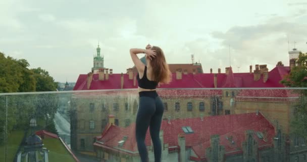 一个喜欢运动的女孩站在那里看着城市。她正在理直头发,扭着屁股.这个女孩穿着紧身裤和太阳镜。背景中房屋的屋顶。4K — 图库视频影像