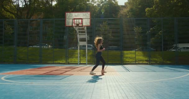 Sportowa dziewczyna robi przysiady z wyskakiwaniem i skakaniem na różnych nogach. Ćwiczy na boisku do kosza. Ubiera się w strój sportowy. 4K — Wideo stockowe