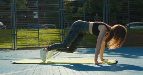 Sport flicka gör övningen "bergsklättrare". Hon tränar på yogamattan på basketplanen. Hon klär sig i en sportuniform. 4K — Stockvideo