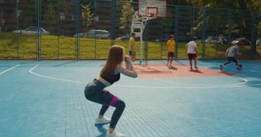 Spor kızı elastik bir bantla çömelme hareketi yapıyor. Kenara adım atarak çömelme hareketleri yapıyor. İnsanlar arka planda hokey oynuyorlar. Kamera bir daire içinde hareket eder. 4K