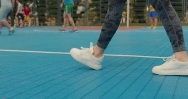 Kızların bacaklarına yakın çekim. Bir kız basketbol sahasında yürüyor. İnsanlar arka planda hokey oynuyorlar. 4K