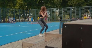 Spor kızı basamaklarda zıplayarak çömelme hareketleri yapıyor. Saçları rüzgarda uçuşuyor. Spor üniforması giyiyor. Kamera onun etrafında hareket ediyor. İnsanlar arka planda hokey oynuyor.