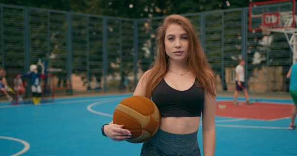 Disparos de cerca. Retrato de una chica deportiva. La chica está mirando a la cámara. Ella sostiene una pelota de baloncesto. La gente está jugando al hockey en el fondo. 4K — Vídeo de stock