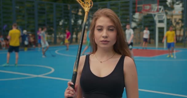 Close-up shooting. Portrait of a sports girl. The girl is looking at the camera and lifting the hockey stick on her shoulder. Her hair is fluttering in the wind. People are playing hockey in the — Stock Video