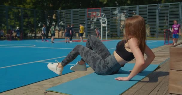 Дівчина потискає свою пресу під час виконання велотренувань. Вона сидить на килимку для йоги на сцені. Шейс у спортивній формі. Люди грають в хокей на задньому плані. Камера рухається — стокове відео