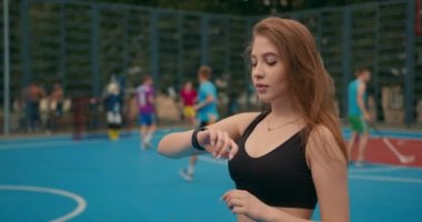 Yakın plan çekim. Spor kızı portresi. Kız akıllı seğirmesindeki eğitim bilgilerine bakıyor. Saçları rüzgarda uçuşuyor. İnsanlar arka planda hokey oynuyorlar. Kamera.