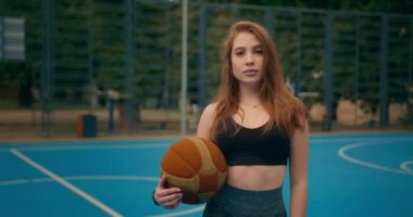 Yakın plan çekim. Spor kızı portresi. Kız, kameraya bakıyor. Elinde basketbol topu tutuyor. İnsanlar arka planda hokey oynuyorlar. Kamera bir daire içinde hareket ediyor. 4K