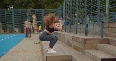 Orta boy shot. Spor kızı basamaklarda zıplayarak çömelme hareketleri yapıyor. Saçları rüzgarda uçuşuyor. Spor üniforması giyiyor. Kamera onun etrafında hareket ediyor. İnsanlar hokey oynuyor.