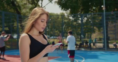 Yakın plan çekim. Sportif bir kız akıllı telefonuyla mesaj atıyor. Saçları rüzgarda uçuşuyor. Spor üniforması giyiyor. İnsanlar arka planda hokey oynuyorlar. 4K