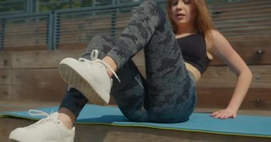 Yakın plan çekim. Bir kız bisiklet egzersizi yaparken presini sallıyor. Bir basamakta yoga minderinde oturuyor. Spor üniforması giyiyor. İnsanlar arka planda hokey oynuyor.