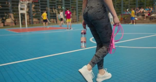 Närbild skytte. Sporttjejen går på basketplanen. Hon håller i ett hopprep och en flaska vatten. Hon bär leggings. Folk spelar hockey i bakgrunden. 4K — Stockvideo