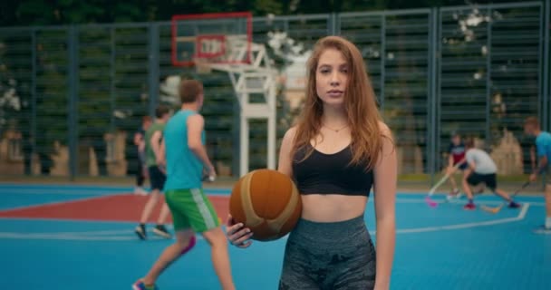 Strzelanina z bliska. Portret dziewczyny ze sportu. Dziewczyna patrzy w kamerę i pozuje. Trzyma koszykówkę. Ludzie grają w hokeja w tle. Kamera porusza się w kółko. — Wideo stockowe