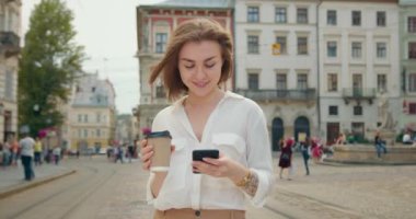 Kız telefonda mesajlaşıyordu. O ayakta ve gülümsüyor. Elinde bir fincan kahve tutuyor. Güneş parlıyor ve insanlar arka planda yürüyorlar. 4K