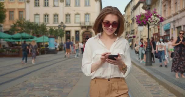 Una chica sonriente está caminando y enviando mensajes de texto en su teléfono inteligente. Lleva gafas de sol. La gente camina en el fondo. 4K — Vídeos de Stock