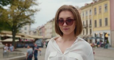 Yakın plan çekim. Portre çekimi. Güneş gözlüklü bir kız kameraya bakıyor. O gülümsüyor. İnsanlar arka planda yürüyorlar. 4K