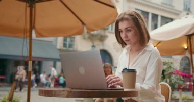 Kız bir kafede verandada oturuyor. Laptopuyla uğraşıyor. Masanın üzerinde bir fincan kahve var. İnsanlar arka planda dinleniyor. 4K
