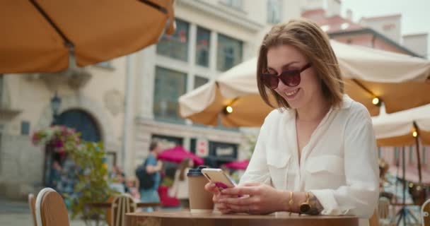 La chica sonriente está sentada en una mesa en un café y mandando mensajes de texto por teléfono. Una taza de café está sobre la mesa. Lleva gafas de sol. La plaza central de la ciudad en el fondo. 4K — Vídeos de Stock