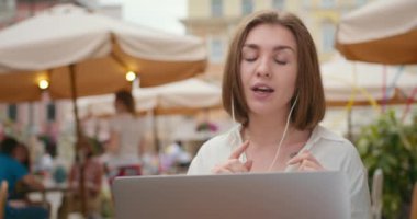 Yakın plan çekim. Kız bir kafede oturuyor ve bir video konferansı düzenliyor. Kulaklıklarıyla konuşuyor. İnsanlar arka planda yürüyorlar. 4K