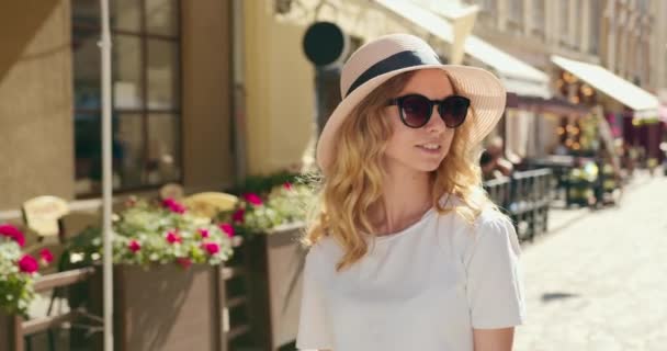 Disparo de retratos. Una chica está caminando por la ciudad. Ella está mirando lo que está pasando a su alrededor. Lleva sombrero y gafas de sol. 4K — Vídeo de stock