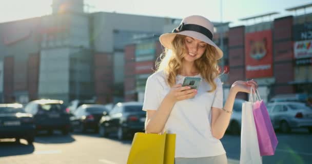 Een lachend meisje sms 't op haar smartphone. Ze heeft veel boodschappentassen in haar handen. Winkelcentrum op de achtergrond. 4K — Stockvideo