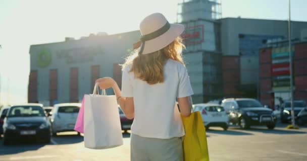 A disparar por trás. A menina está procurando um carro no estacionamento. Ela tem um monte de sacos de compras nas mãos. Centro comercial em segundo plano. 4K — Vídeo de Stock