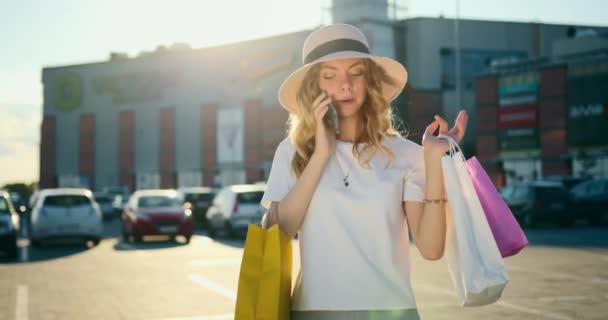 Девушка покидает торговый центр с сумками. Она разговаривает по телефону и улыбается. Солнце светит, а торговый центр находится на заднем плане. 4K — стоковое видео
