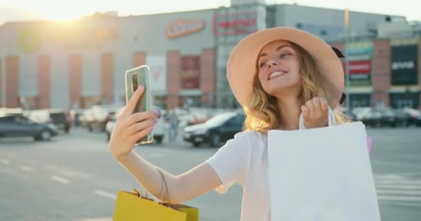 Uma rapariga está a tirar uma selfie no seu smartphone. Ela está sorrindo e segurando os sacos de compras em suas mãos. Centro comercial em segundo plano. 4K — Vídeo de Stock