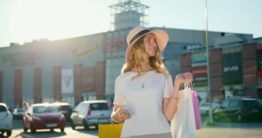 Kız alışveriş merkezinden alışveriş çantalarıyla ayrılıyor. Gülümsüyor ve yürüyor. Şapka takıyor. Güneş parlıyor ve alışveriş merkezi arka planda. 4K