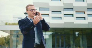 İş adamı telefonda duygu sömürüsü yapıyor. Takım elbise ve gözlük takıyor. İş merkezi arka planda. 4K
