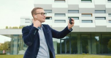 Bir iş adamı iş merkezinin yakınında yürüyor ve telefonda video bağlantısıyla duygusal olarak konuşuyor. Kulaklıkla konuşuyor. Takım elbise ve gözlük takıyor. 4K