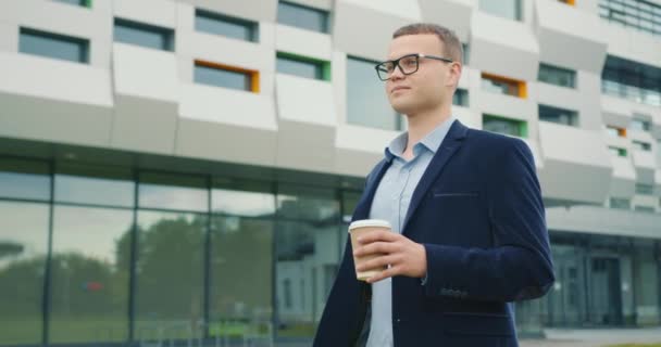 stock video A businessman is walking with a cup of coffee near the business center. He is smiling. He is wearing a suit and glasses. 4K