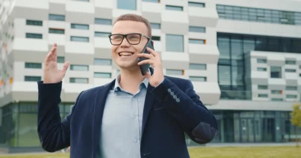 The businessman is going and talking emotionally on the phone. Hes wearing a suit and glasses. The business center is in the background. 4K — Stock Video