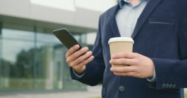 İş merkezinin dışında elinde bir fincan kahveyle bir işadamı duruyor. Akıllı telefonuyla mesaj yazıyormuş. Takım elbise ve gözlük takıyor. Yakın plan çekim. 4K