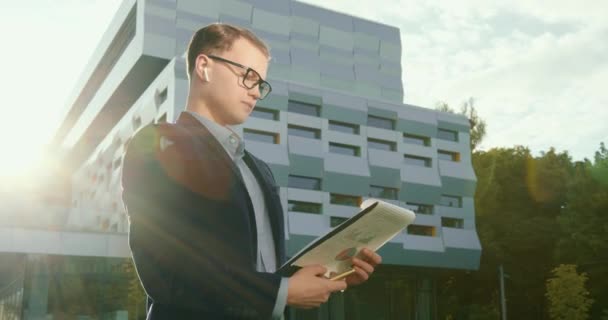 Der Geschäftsmann blättert in Dokumenten und macht sich mit einem Stift Notizen. Er trägt Kopfhörer. Er trägt Anzug und Brille. Business Center im Hintergrund. 4K — Stockvideo