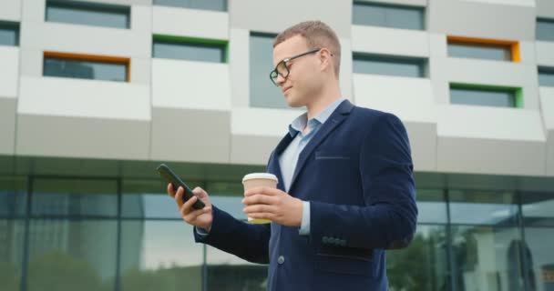 Un om de afaceri stă în fața centrului de afaceri cu o ceașcă de cafea în mână. Îşi trimite mesaje pe smartphone. Poartă un costum şi ochelari. 4K — Videoclip de stoc
