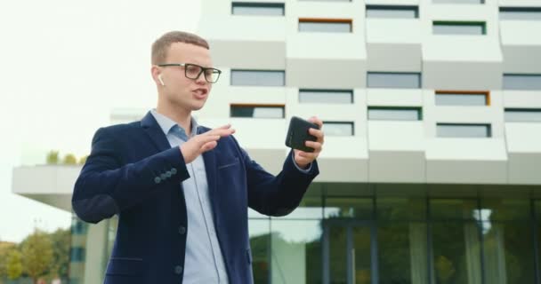 Um homem de negócios está andando perto do centro de negócios e descontente falando via link de vídeo no telefone. Ele está falando em fones de ouvido. Ele está a usar um fato e óculos. 4K — Vídeo de Stock