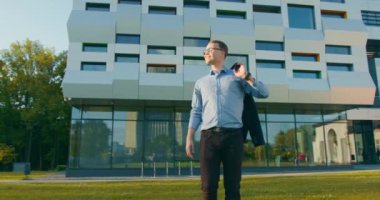 Bir iş adamı yavaşça çimenlerde yürüyor ve güneşin tadını çıkarıyor. Ceketini omzunun üstünde tutuyor. Arka planda iş merkezi var. 4K