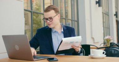 Bir işadamı belgelere bakıyor ve bir dizüstü bilgisayar üzerinde çalışıyor. O, bir kafede bir masada oturuyor. Gülümsüyor. Masanın üzerinde bir fincan kahve var. 4K