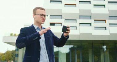 Bir işadamı iş merkezinin yakınında yürüyor ve telefonda video bağlantısıyla hoşnutsuzca konuşuyor. Kulaklıkla konuşuyor. Takım elbise ve gözlük takıyor. 4K