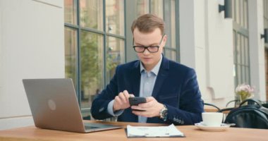 Bir iş adamı bir kafede bir masada oturuyor ve çalışıyor. O, telefonda bilgi arıyor ve dizüstü bilgisayarda çalışıyor. Masada bir fincan çay var. 4K