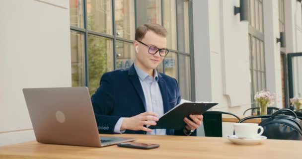 Ein Geschäftsmann blättert in Arbeitspapieren. Er legt die Unterlagen auf den Tisch und nimmt eine Tasse Tee. Es macht ihm Spaß. Arbeit in einem Café. 4K — Stockvideo