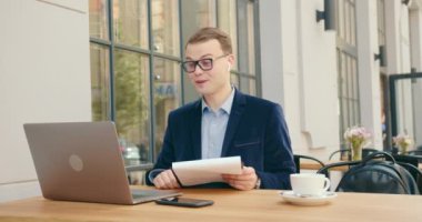 Bir iş adamı, iş meselelerini bir laptoptaki video görüşmesiyle neşeyle tartışıyor. Kulaklıkla konuşuyor. O bir kafede oturuyor ve masanın üzerinde bir fincan çay içiyor. 4K