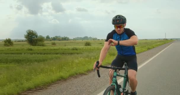 Велосипедист дивиться на дані тренувань на трекер фітнесу. Він починає збільшувати швидкість їзди. Навчання на велосипеді. На задньому плані зелене поле. 4-кілометровий — стокове відео